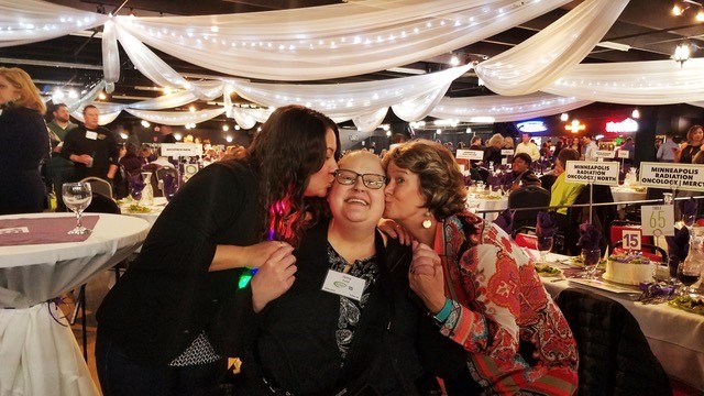 Two female MRO staffers giving a smiling female patient a kiss on the cheek at fundraiser event.